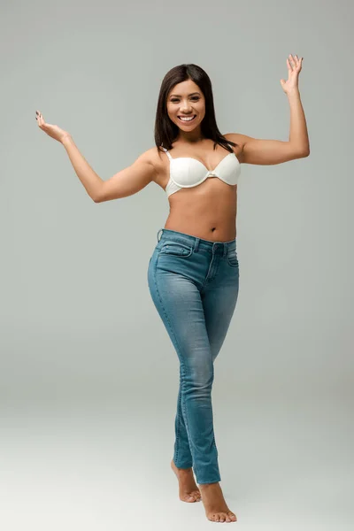 Übergewichtige und glückliche Afroamerikanerin in Jeans und BH steht auf grau — Stockfoto