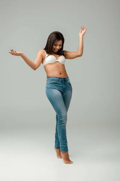 Толстая и счастливая африканская американка в джинсах и лифчике, стоящая на сером — стоковое фото