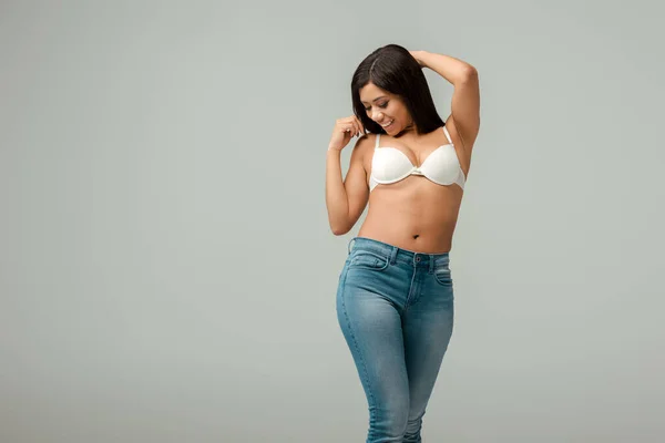 Übergewichtige und glückliche Afroamerikanerin in Jeans und BH steht isoliert auf grau — Stockfoto