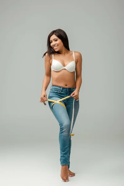 Niña afroamericana sonriente y con sobrepeso en jeans y sujetador midiendo el muslo en gris - foto de stock