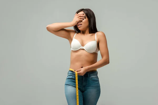 Sobrepeso menina afro-americana em jeans e sutiã medindo cintura e cobrindo olhos isolados em cinza — Fotografia de Stock