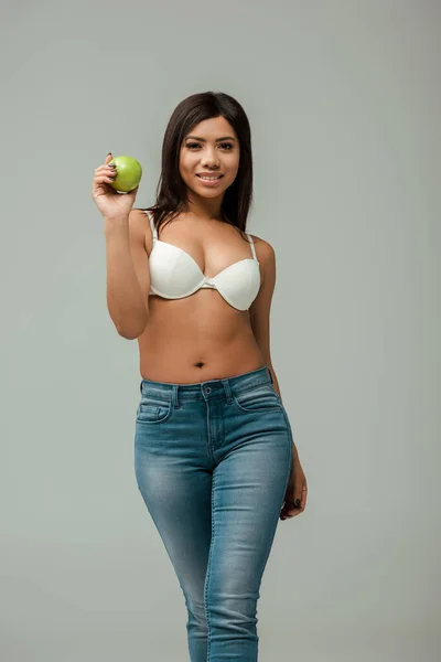 Happy and overweight african american woman in jeans and bra holding apple isolated on grey — Stock Photo