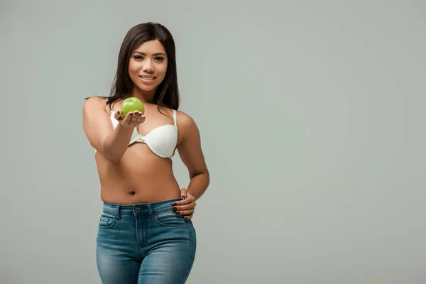 Cheerful and overweight african american woman in jeans and bra holding apple isolated on grey — Stock Photo