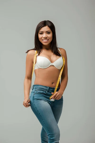 Happy and overweight african american girl in jeans and bra holding measuring tape isolated on grey — Stock Photo