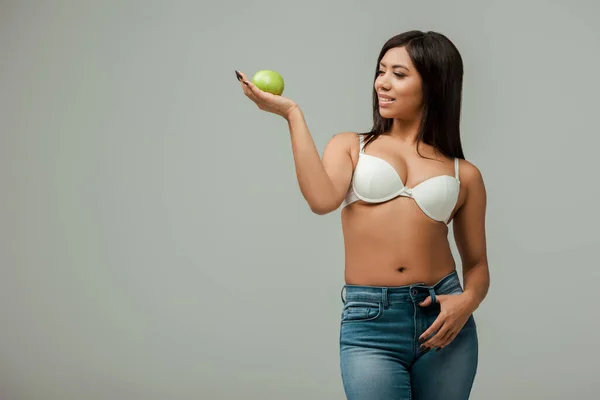 Cheerful and overweight african american girl in jeans and bra holding apple isolated on grey — Stock Photo