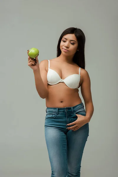 Overweight african american girl in jeans and bra looking at apple isolated on grey — Stock Photo