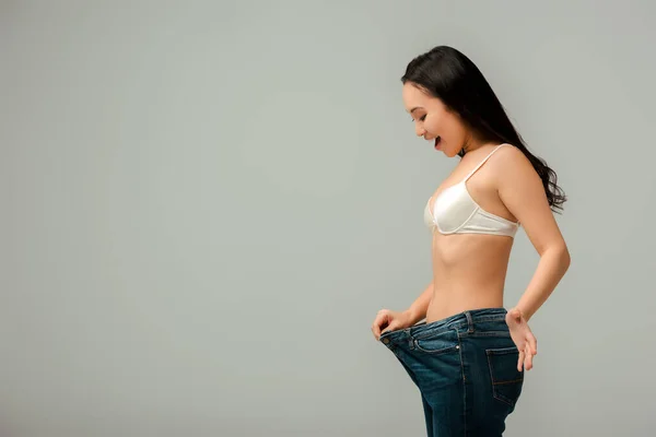 Aufgeregtes asiatisches Mädchen berührt übergroße Jeans isoliert auf grau — Stockfoto