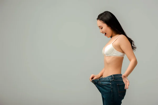 Excited asian girl touching oversized jeans isolated on grey — Stock Photo