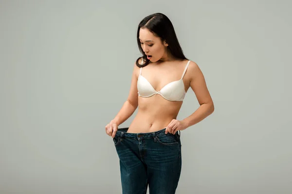 Sorprendido asiático chica tocando sobredimensionado jeans aislado en gris - foto de stock