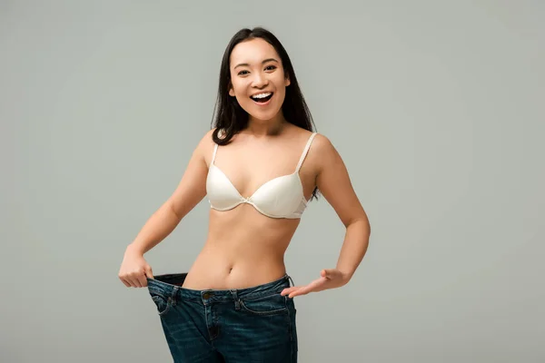 Cheerful asian girl pointing with hand at oversized jeans isolated on grey — Stock Photo