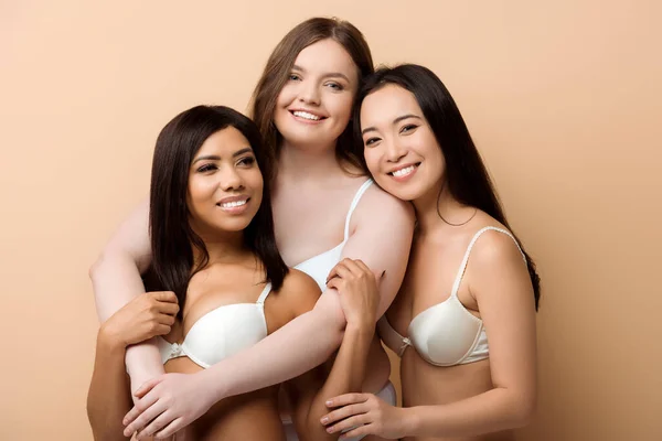 Smiling plus size multicultural women in bras hugging isolated on beige — Stock Photo