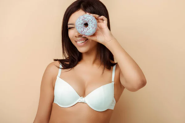 Glücklich afrikanisch-amerikanische Mädchen in BH hält Donut in Augennähe isoliert auf beige, Körper positives Konzept — Stockfoto