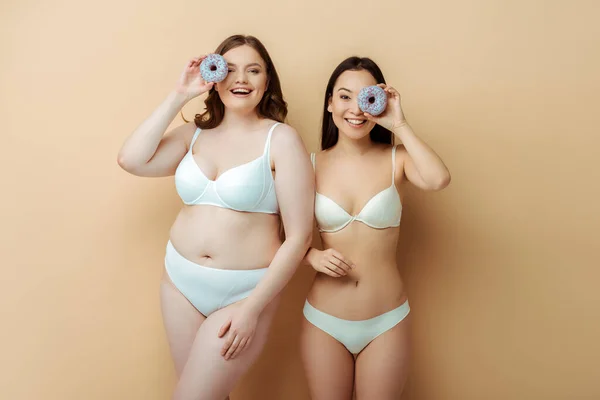 Happy plus size woman and asian girl in underwear holding doughnuts near eyes isolated on beige, body positive concept — Stock Photo