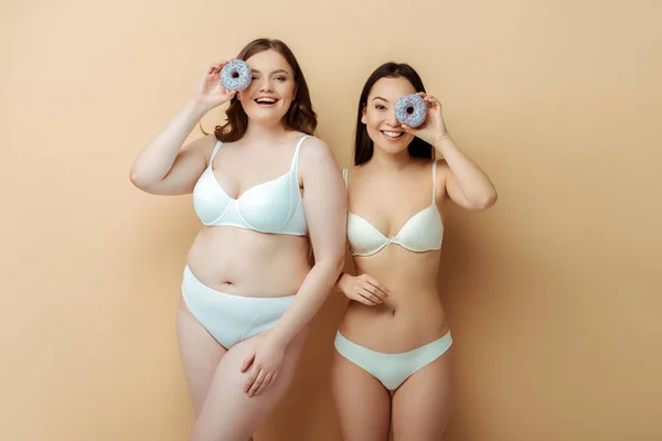 Felice plus size donna e ragazza asiatica in biancheria intima in possesso di ciambelle vicino agli occhi isolati su beige, concetto positivo per il corpo — Foto stock