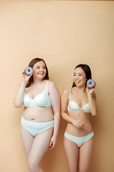 Feliz más tamaño mujer y asiático chica en ropa interior celebración donas aislado en beige, cuerpo positivo concepto - foto de stock