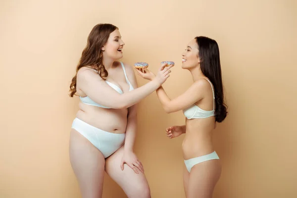Cheerful plus size woman and asian girl in underwear holding doughnuts isolated on beige, body positive concept — Stock Photo