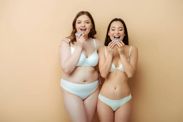 Happy plus size woman and asian girl in underwear eating doughnuts isolated on beige, body positive concept — Stock Photo