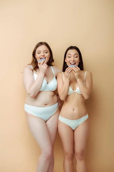 Счастливая плюс размер женщина и веселая азиатская девушка в нижнем белье едят пончики изолированы на бежевый, тело положительное понятие — стоковое фото