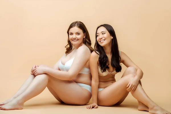 Allegra donna in sovrappeso e felice ragazza asiatica in biancheria intima seduta sul beige, concetto positivo per il corpo — Foto stock