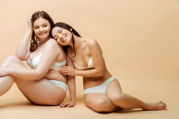 Cheerful asian girl sitting with overweight woman in underwear on beige, body positive concept — Stock Photo