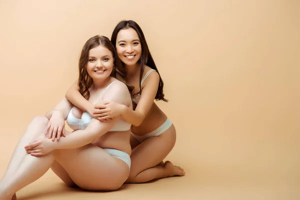 Fröhliches asiatisches Mädchen umarmt übergewichtige Frau in Unterwäsche, während sie auf beigem, körperpositivem Konzept sitzt — Stockfoto