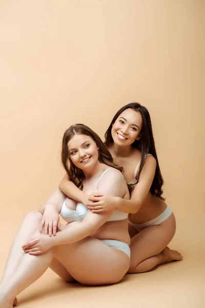 Happy asian girl hugging overweight woman in underwear while sitting on beige, body positive concept — Stock Photo