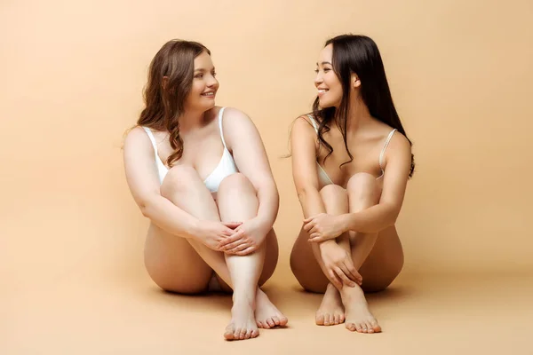 Cheerful plus size woman and smiling asian girl in lingerie sitting on beige, body positive concept — Stock Photo