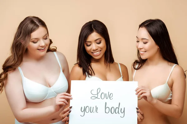 Happy and overweight multicultural women in bras holding placard with love your body lettering isolated on beige, body positive concept — Stock Photo