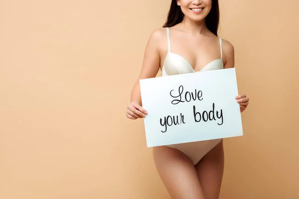 Vista ritagliata di allegra ragazza tenendo cartello con amore il tuo corpo lettering isolato su beige — Foto stock