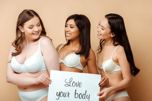 Mulheres multiculturais felizes em sutiãs segurando cartaz com amor seu corpo lettering isolado em bege, corpo conceito positivo — Fotografia de Stock