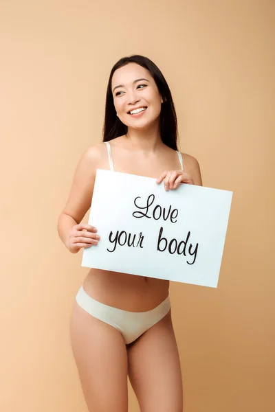 Alegre asiático chica holding cartel con amor su cuerpo lettering aislado en beige - foto de stock