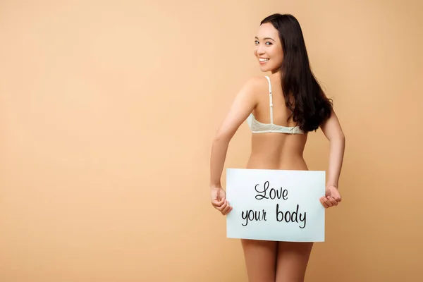 Feliz asiático chica holding cartel con amor su cuerpo letras aislado en beige - foto de stock