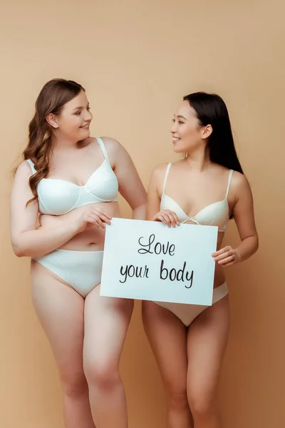 Cheerful multicultural girls in underwear holding placard with love your body lettering isolated on beige, body positive concept — Stock Photo