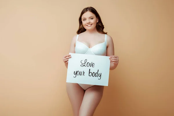 Felice plus size ragazza tenendo cartello con amore il tuo corpo lettering isolato su beige, concetto positivo corpo — Foto stock