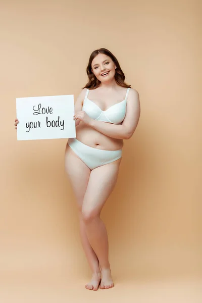 Plus size donna sorridente, guardando la fotocamera e mostrando cartello con amore il tuo corpo lettering sul beige — Foto stock