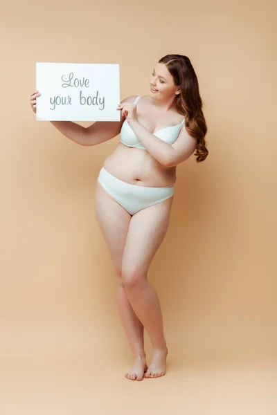 Plus size donna sorridente e tenendo cartello con amore l'iscrizione del corpo su sfondo beige — Foto stock