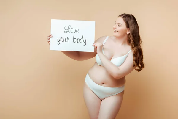 Plus size donna sorridente e mostrando cartello con amore il tuo corpo lettering sul beige — Foto stock