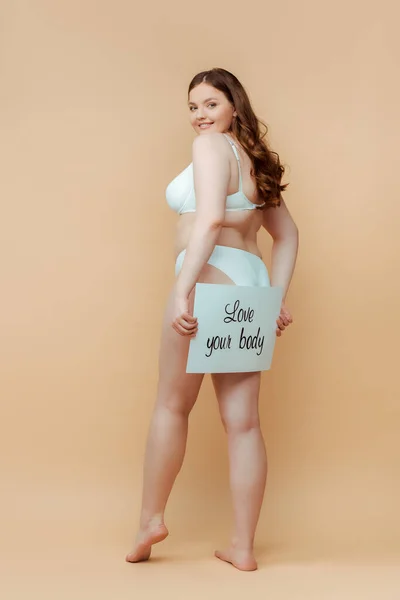 Plus Size Frau in Dessous lächelt und zeigt Plakat mit Love Your Body Schriftzug auf beigem Hintergrund — Stockfoto