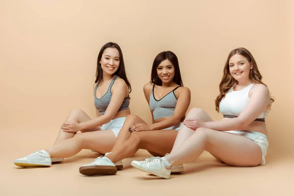 Multiethnic sportswomen sitting, smiling and looking at camera on beige — Stock Photo