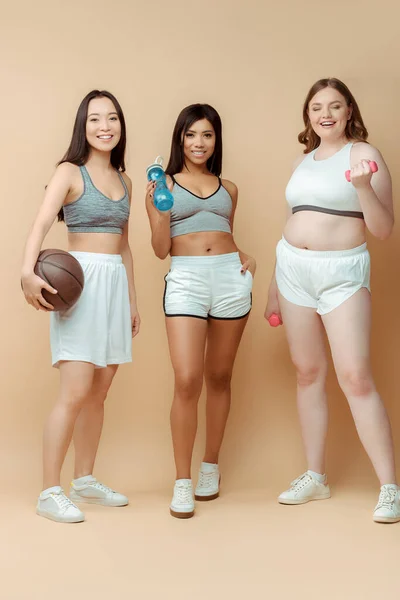Multiethnische Sportlerinnen mit Ball und Sportflasche lächelnd in der Nähe von plus size Sportlerinnen mit Hanteln auf beige — Stockfoto