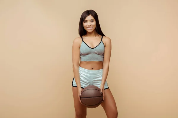 Front view of beautiful african american sportswoman smiling, holding ball and looking at camera isolated on beige — Stock Photo