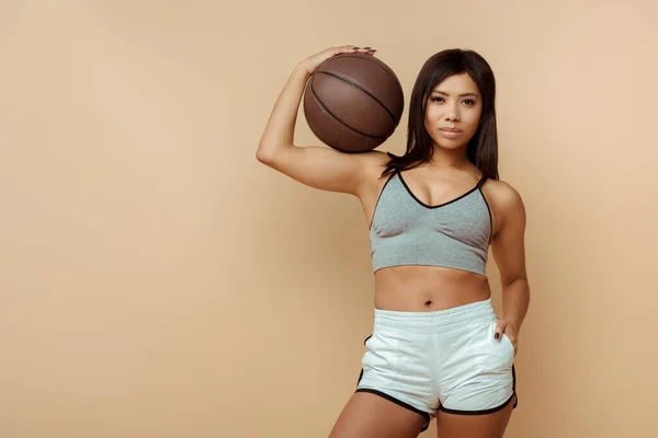 African american sportswoman with hand in pocket and ball looking at camera isolated on beige — Stock Photo