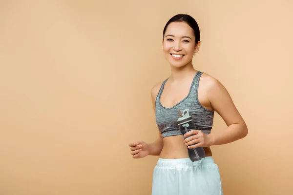 Asiatische Sportlerin mit Sportflasche lächelt und blickt isoliert in die Kamera auf Beige — Stockfoto