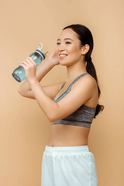 Азиатская спортсменка улыбается со спортивной бутылкой, изолированной на бежевый цвет — стоковое фото