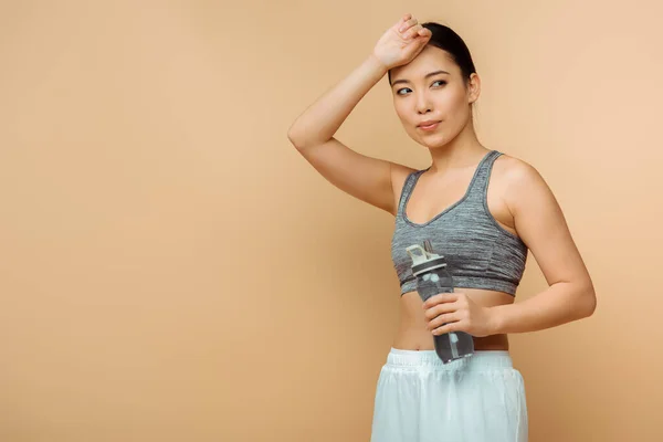Asian sportswoman with sports bottle touching head and looking away on beige — Stock Photo