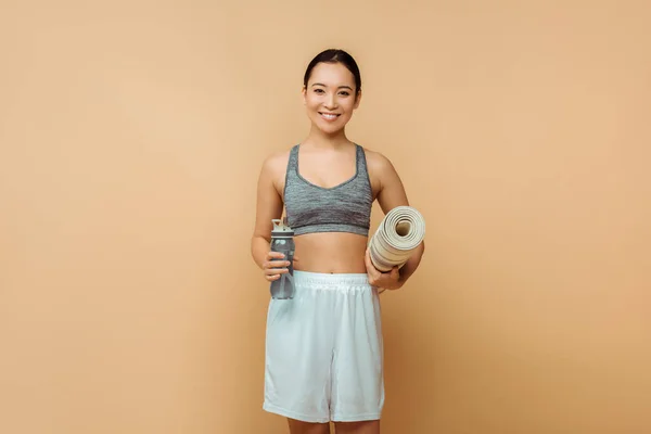 Front view of asian sportswoman smiling, looking at camera and holding sports bottle and fitness mat on beige — Stock Photo