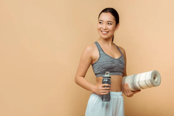 Asiatica sportiva sorridente, guardando altrove e tenendo bottiglia sportiva con tappetino fitness su sfondo beige — Foto stock