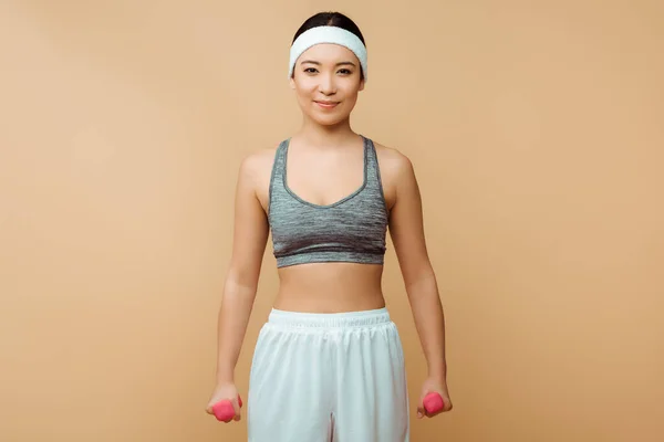 Asian sportswoman with dumbbells smiling and looking at camera on isolated on beige — Stock Photo