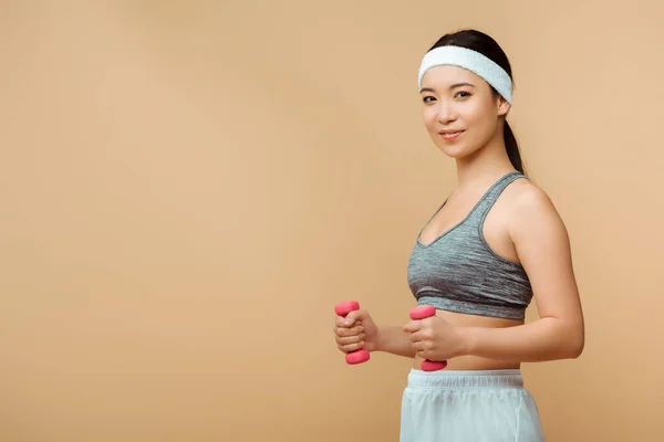 Asiatische Sportlerin mit Hanteln lächelt und blickt isoliert in die Kamera auf beige — Stockfoto