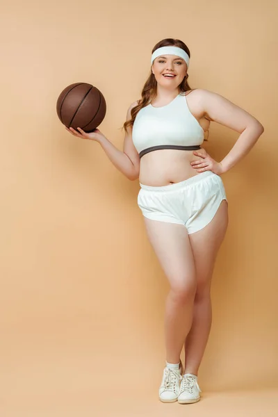 Femme sportive de taille plus avec la main sur la hanche souriant, tenant la balle et regardant la caméra sur fond beige — Photo de stock